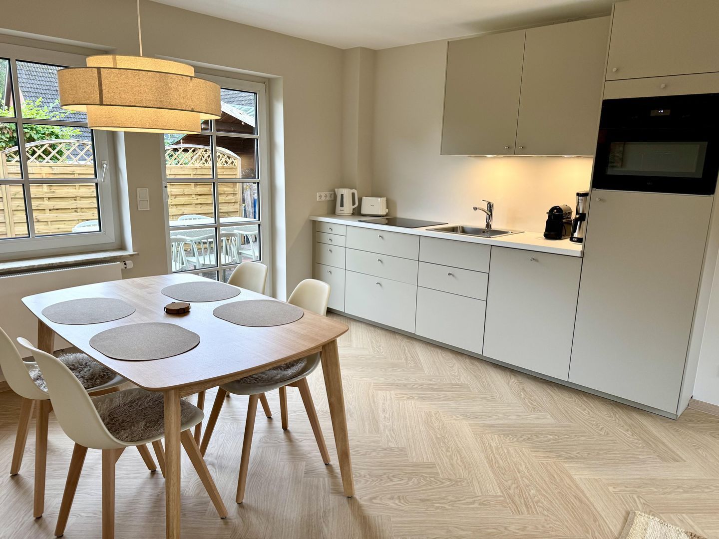 A modern kitchen with a dining area, featuring a wooden table, chairs, a hanging light fixture, and a view of the outdoors through large windows.