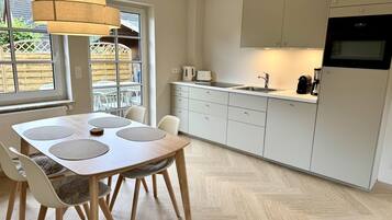 A modern kitchen with a dining area, featuring a wooden table, chairs, a hanging light fixture, and a view of the outdoors through large windows.