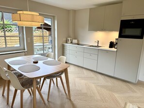 Eine moderne Küche mit Essbereich, ausgestattet mit einem Holztisch, Stühlen, einer Hängeleuchte und Blick ins Freie durch große Fenster.