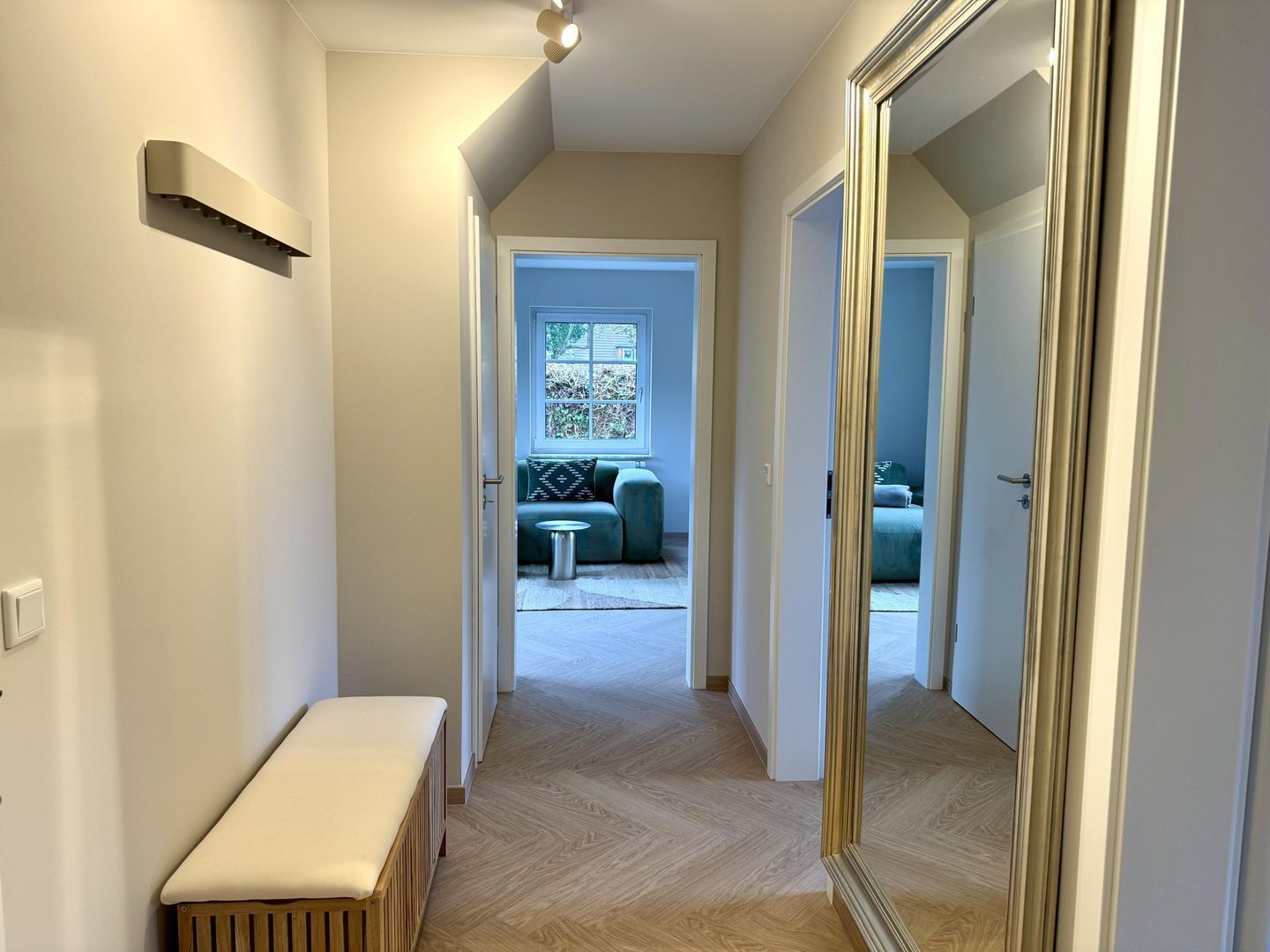 A hallway with a bench, a mirror, and a door leading to a room with a bed and a window.