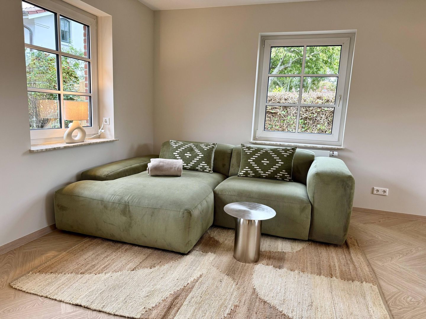 A room with a green sectional sofa, a small round table, a patterned throw pillow, a lamp on a windowsill, and a view of greenery outside.