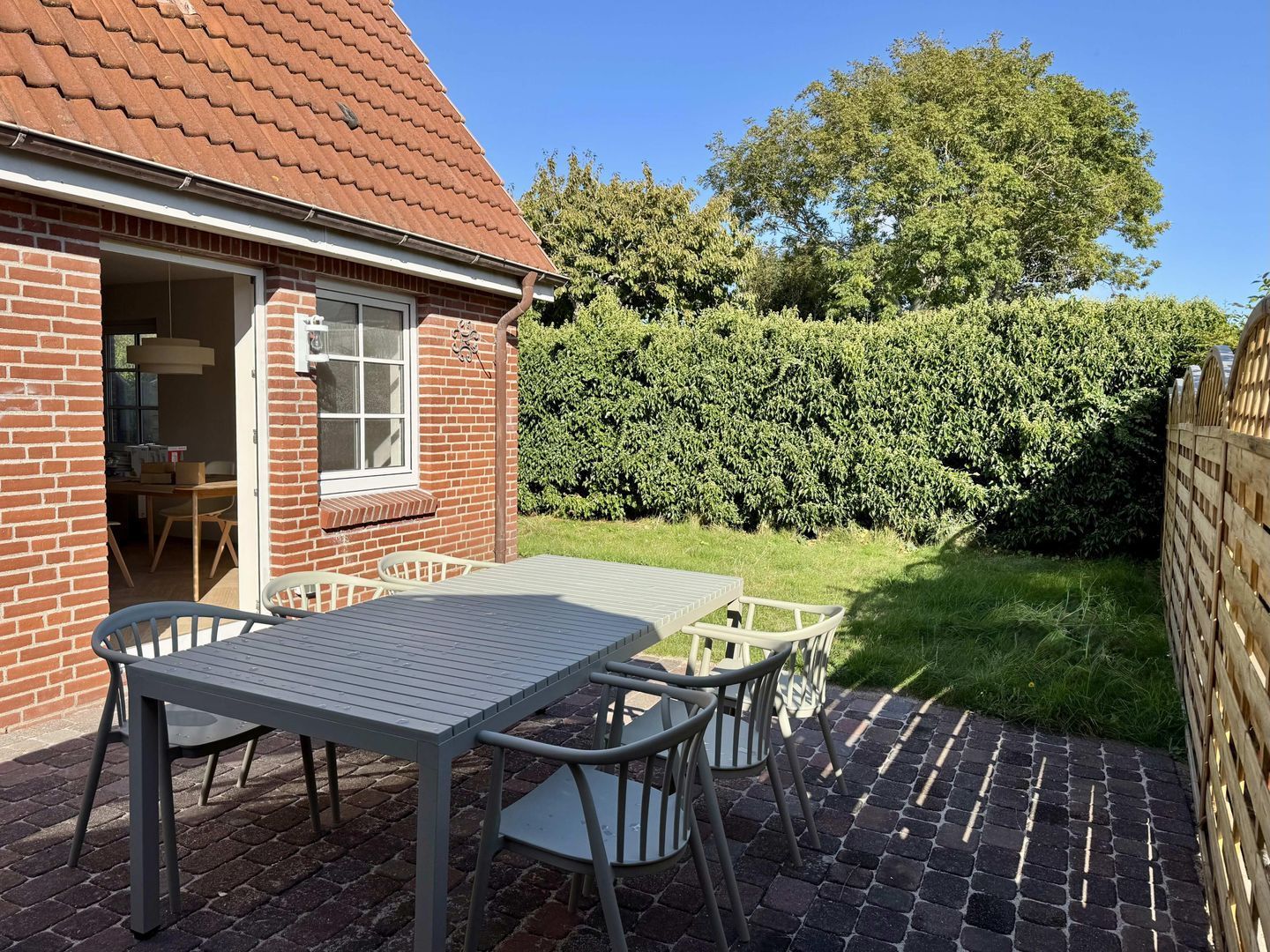 Eine Terrasse mit Tisch und Stühlen, ein Haus mit Ziegelbau und rotem Dach sowie ein Hecken im Hintergrund.