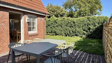 Eine Terrasse mit Tisch und Stühlen, ein Haus mit Ziegelbau und rotem Dach sowie ein Hecken im Hintergrund.