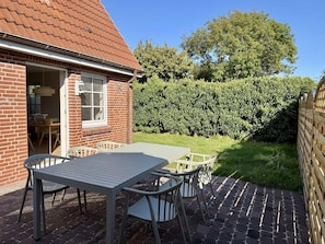 Eine Terrasse mit Tisch und Stühlen, ein Haus mit Ziegelbau und rotem Dach sowie ein Hecken im Hintergrund.
