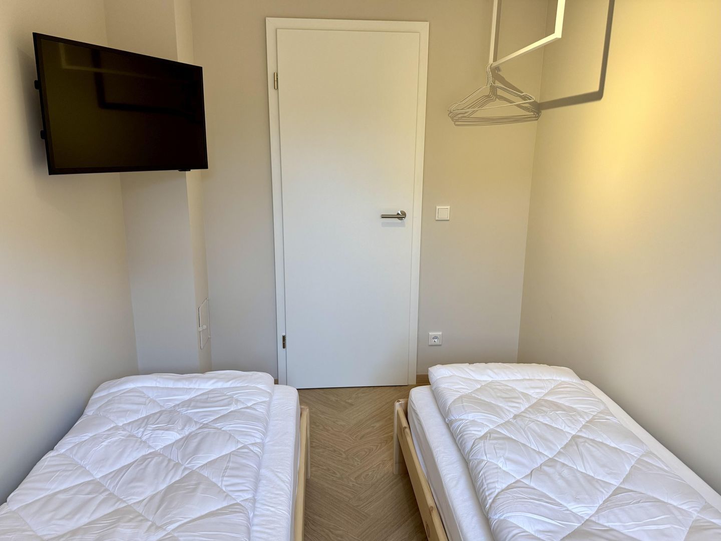 A room with two beds, a door, a wall-mounted TV, and a hanging clothes rack.