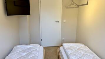 Ein Zimmer mit zwei Betten, einer Tür, einem an der Wand montierten Fernseher und einem Kleiderständer.