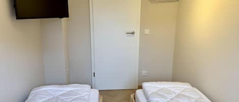 A room with two beds, a door, a wall-mounted TV, and a hanging clothes rack.