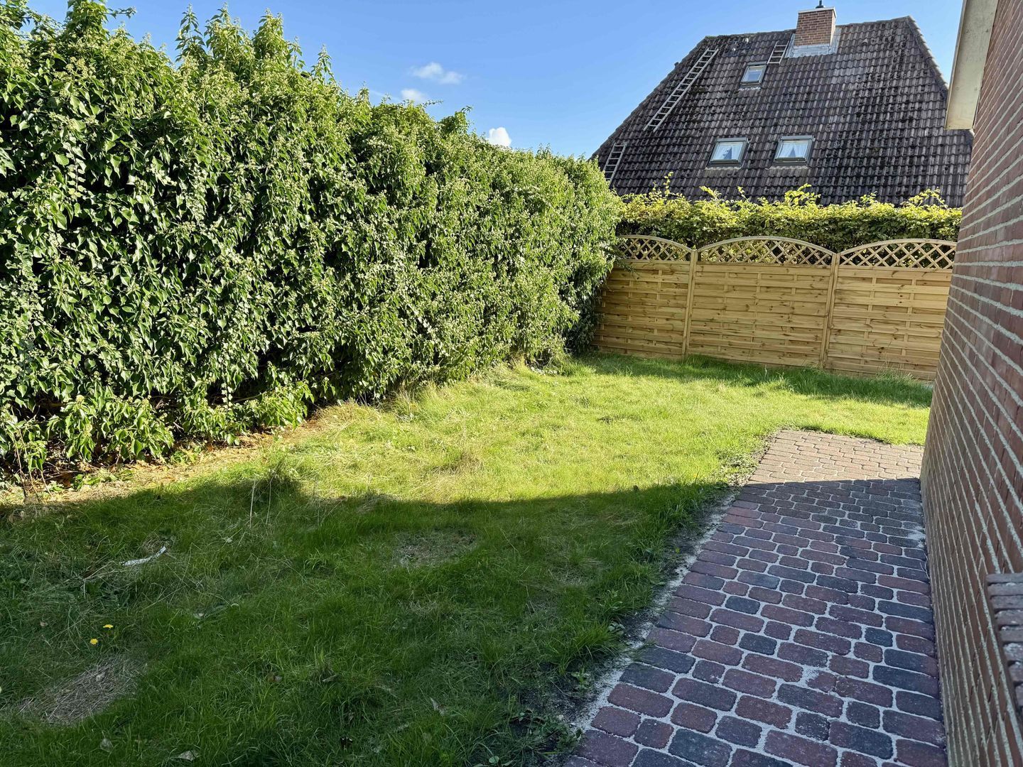 A residential backyard with a grassy lawn, a brick pathway, a wooden fence, and a house with a sloped roof.