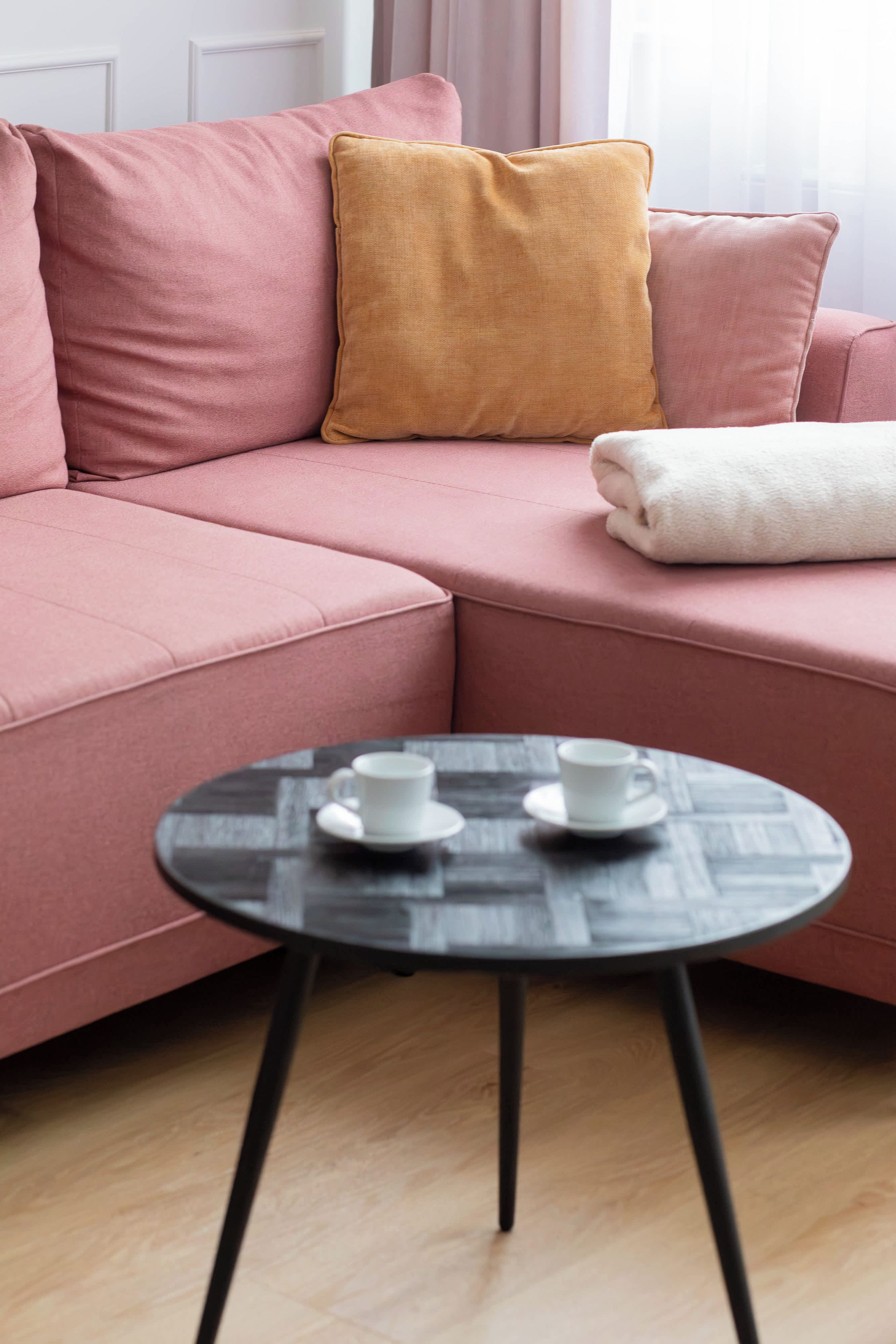 A pink sofa with a mustard-colored pillow and a white towel, a small round table with two white cups, and a wooden floor.