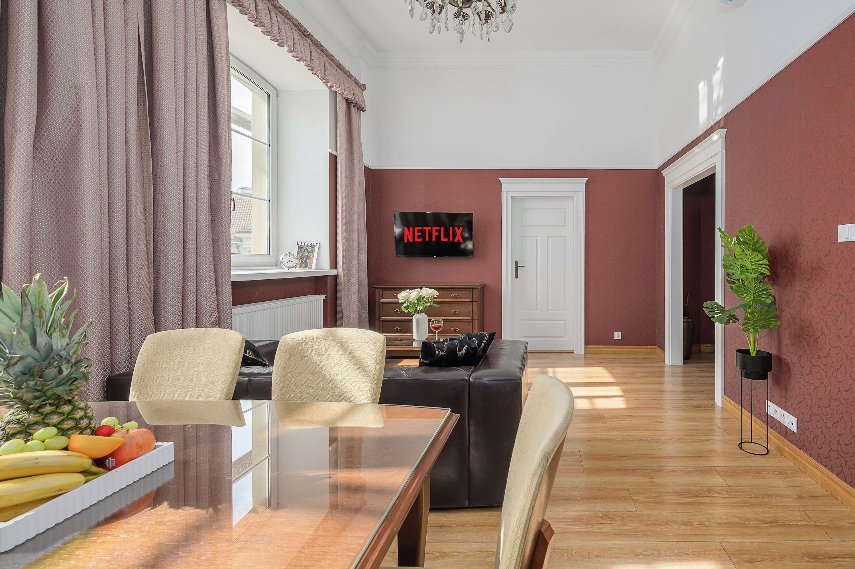A living room with a glass coffee table, two armchairs, a sofa, a TV mounted on the wall, and a door leading to another room.