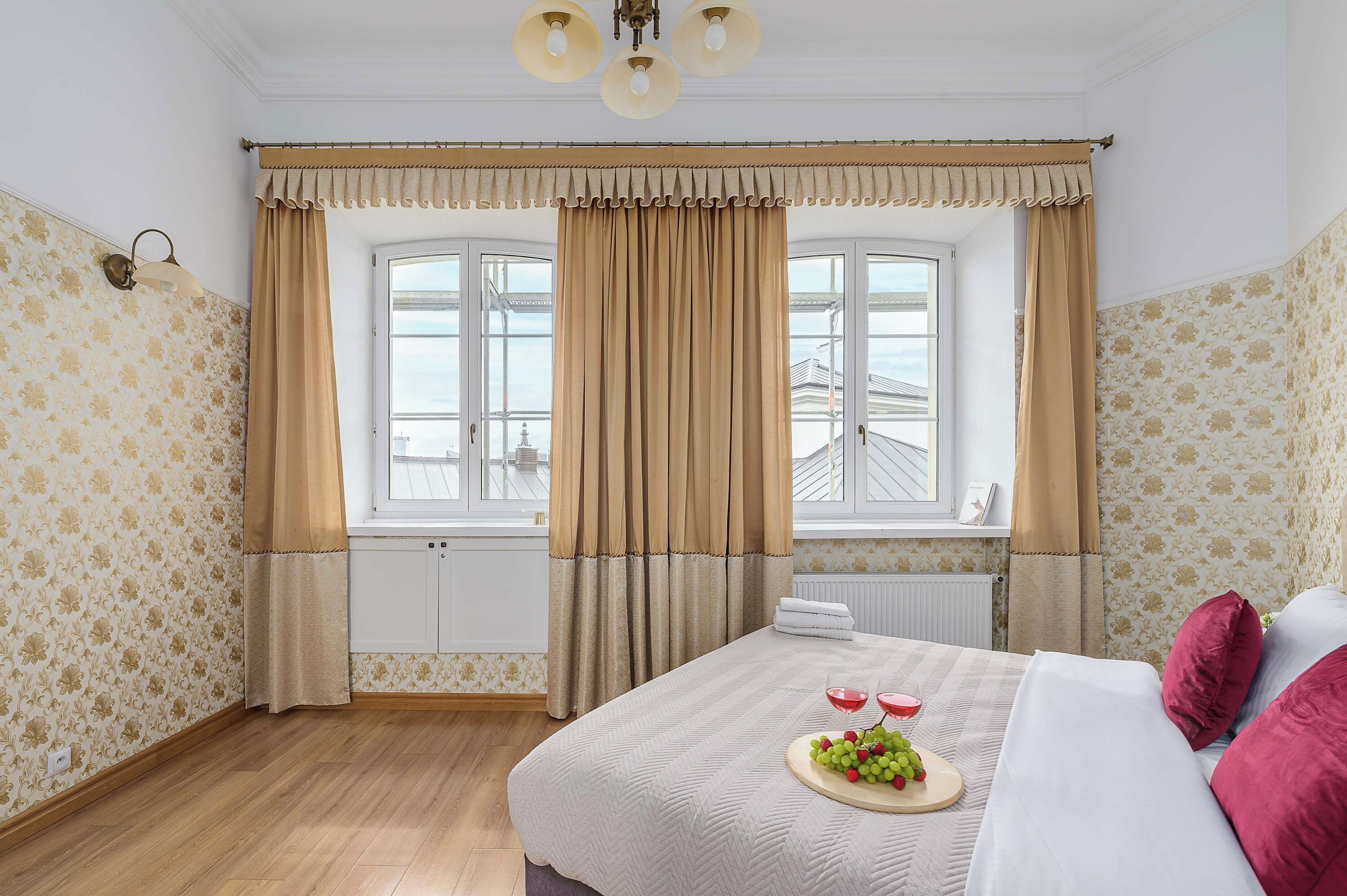 A hotel room with a large bed, a window with curtains, a radiator, and a wall with patterned wallpaper.