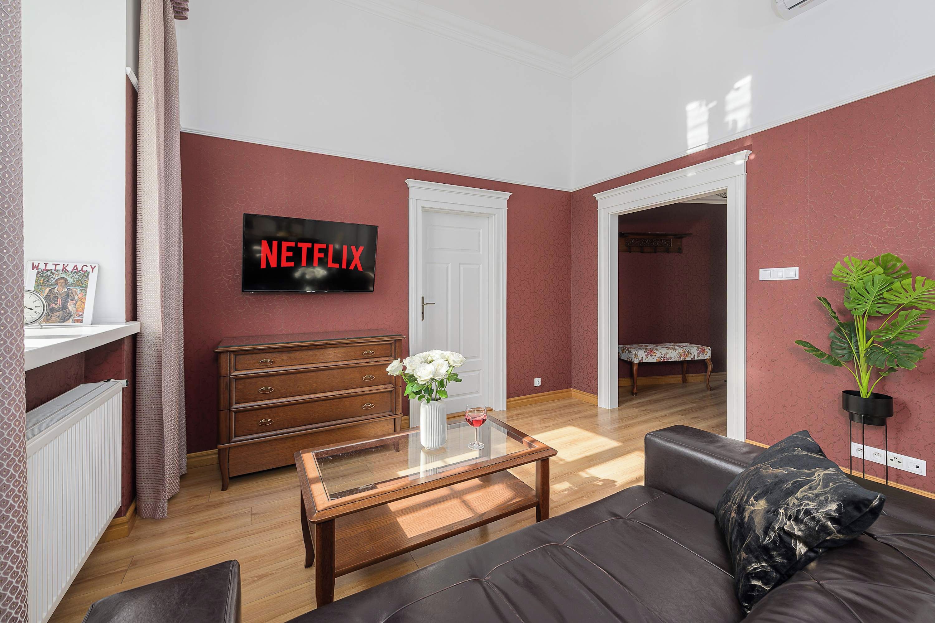 Un salon avec une table basse en bois, un canapé noir, une porte blanche et un téléviseur fixé au mur qui affiche « NETFLIX ».