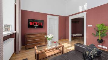 Un salon avec une table basse en bois, un canapé noir, une porte blanche et un téléviseur fixé au mur qui affiche « NETFLIX ».