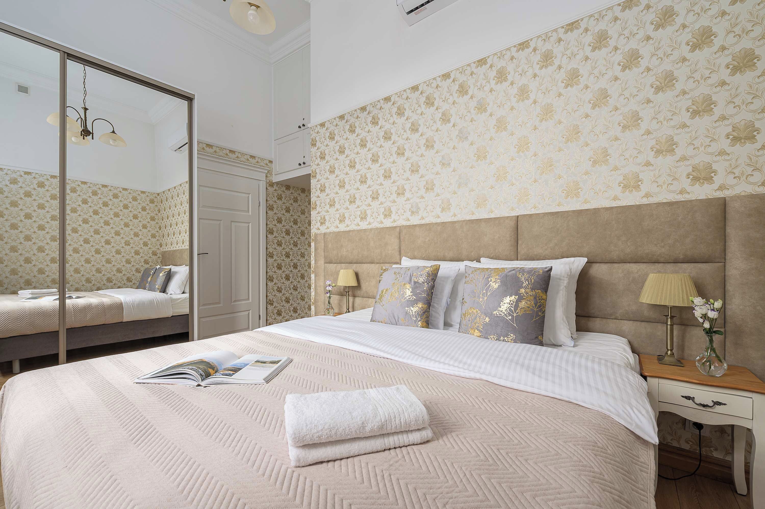 A hotel room with a large bed, a bedside table with a lamp, a mirror, and a wall with floral wallpaper.