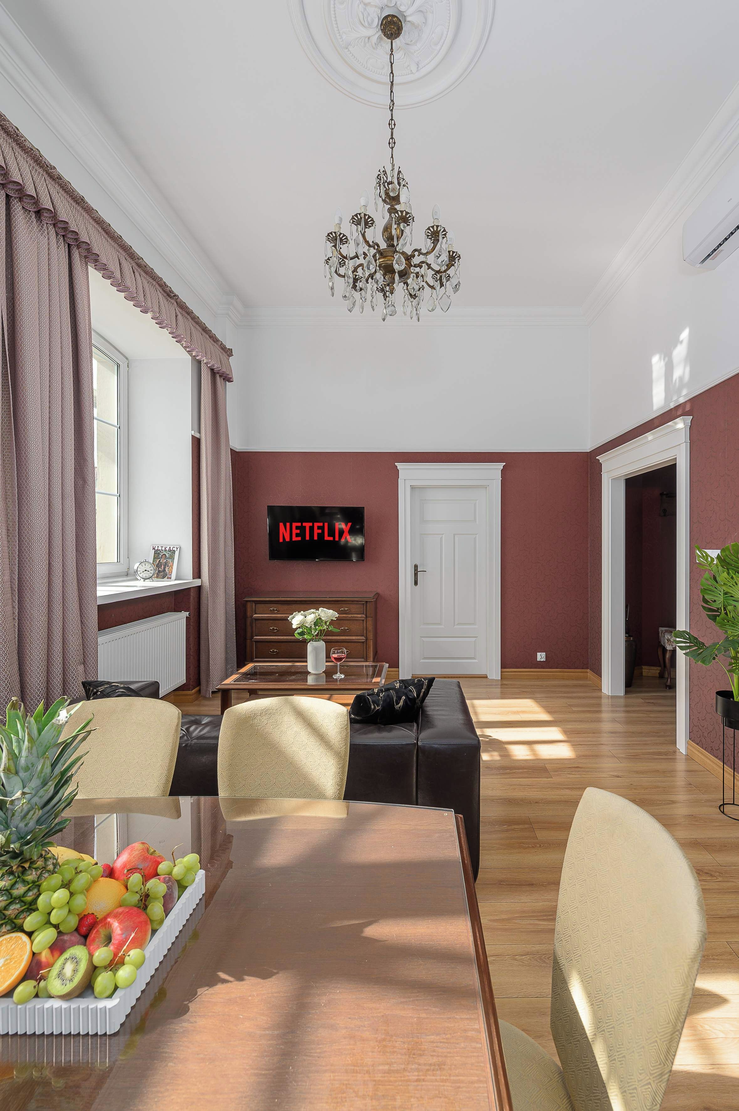 A dining area with a wooden table, chairs, a fruit platter, a television displaying 'NETFLIX', and a chandelier.