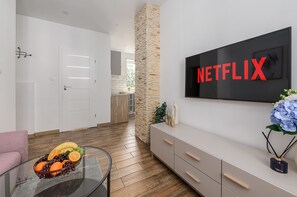A modern living room with a flat-screen TV displaying 'NETFLIX' in red letters, a glass coffee table with fruit, a stone pillar, and a minimalist white cabinet. - Lumina Family 4-room Apart near Metro & Center (Warszawa)