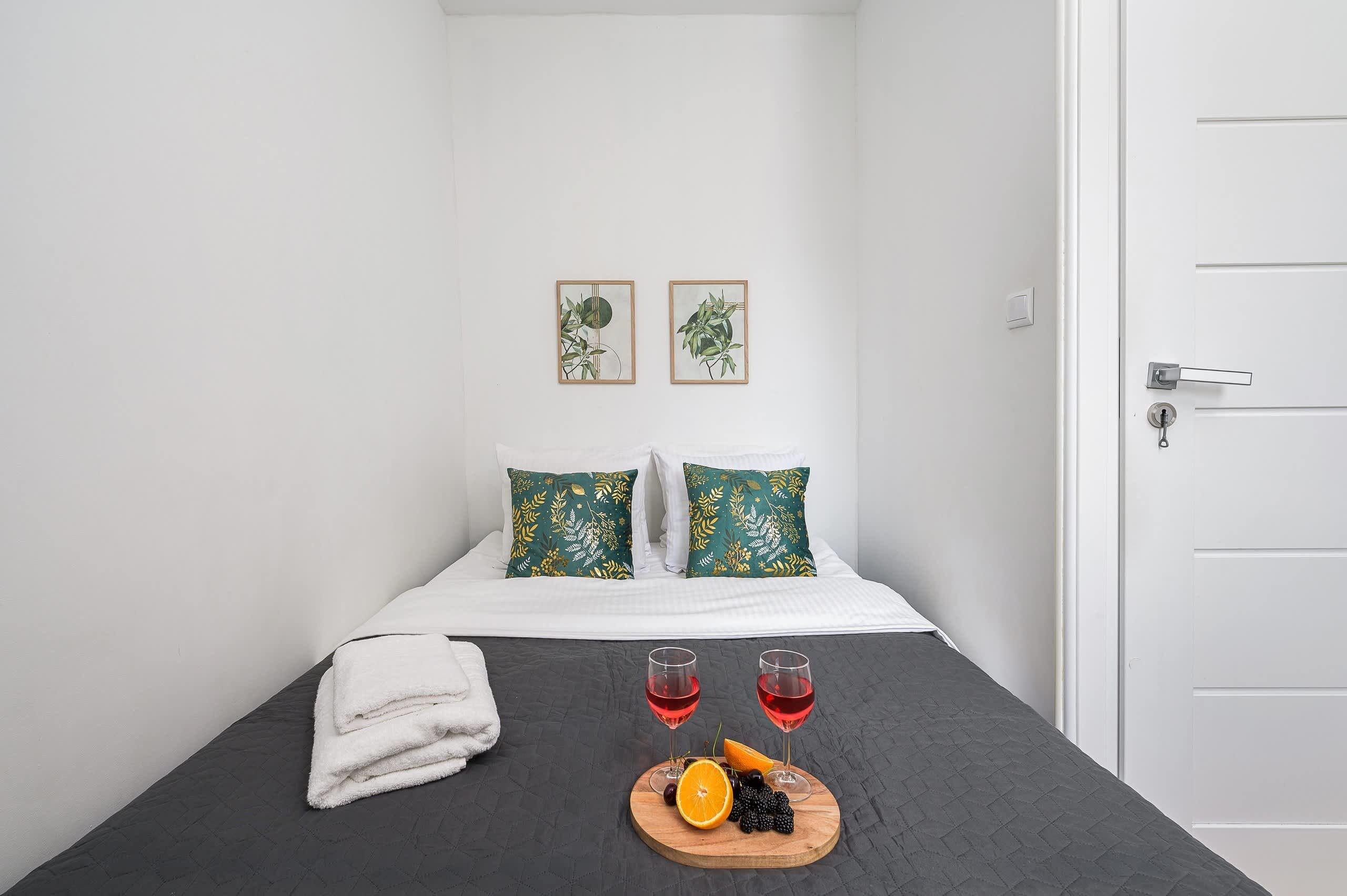 Un lit bien fait, avec une literie blanche et deux coussins à motifs verts, un petit plateau en bois avec des fruits et deux verres d’une boisson rouge, et deux reproductions encadrées de motifs botaniques accrochées au mur.