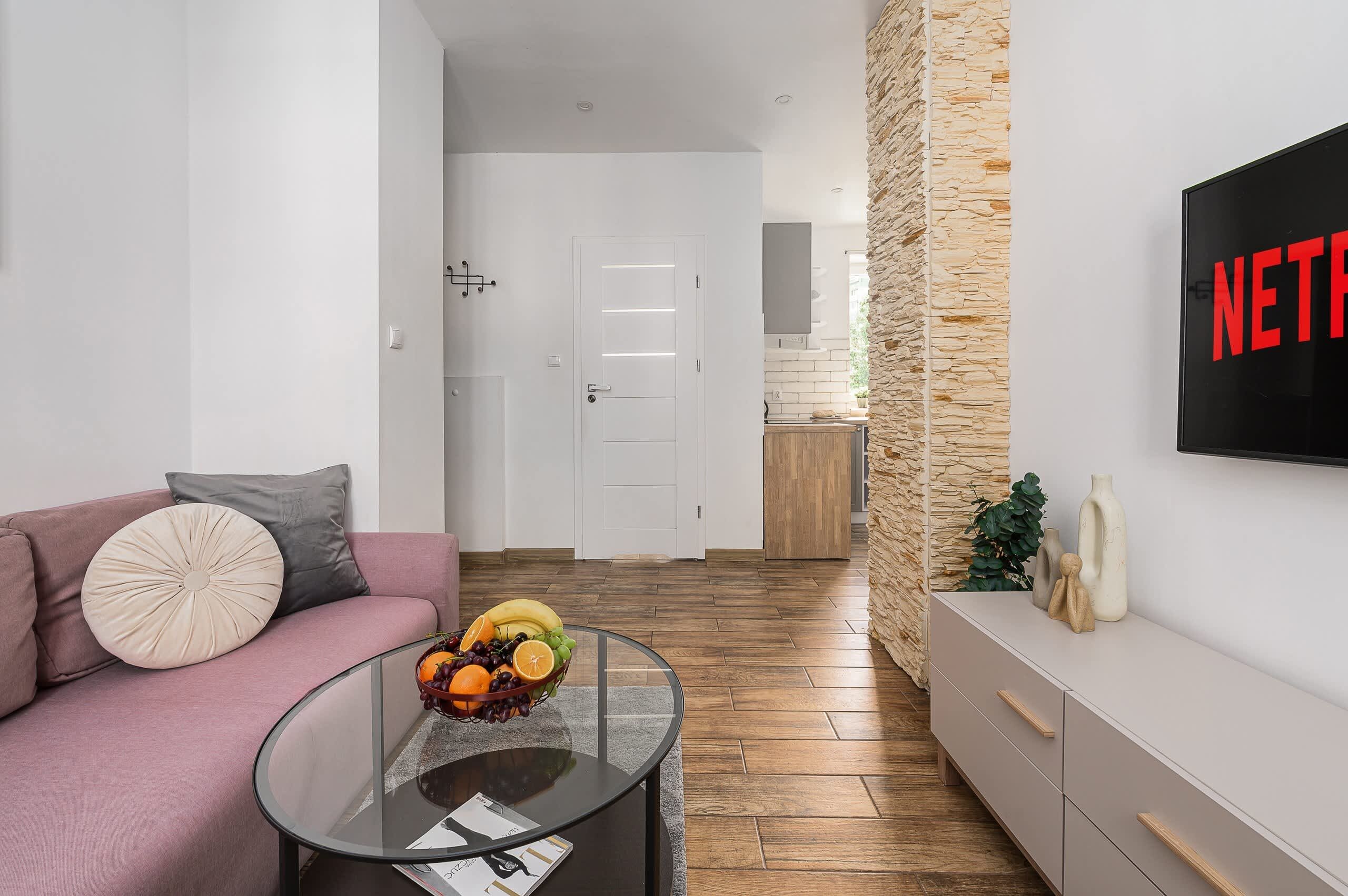 Uma sala de estar moderna com um sofá rosa, uma mesa de centro de vidro com frutas, uma parede de destaque em pedra e uma TV na parede.