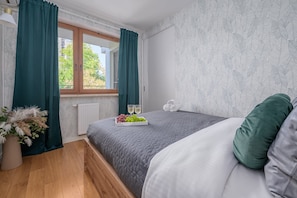 A bedroom with a bed, a window with curtains, a radiator, and a bedside table with a flower arrangement. - Lumina Premium 3-room Apart w/ Parking & Balcony (Warszawa)