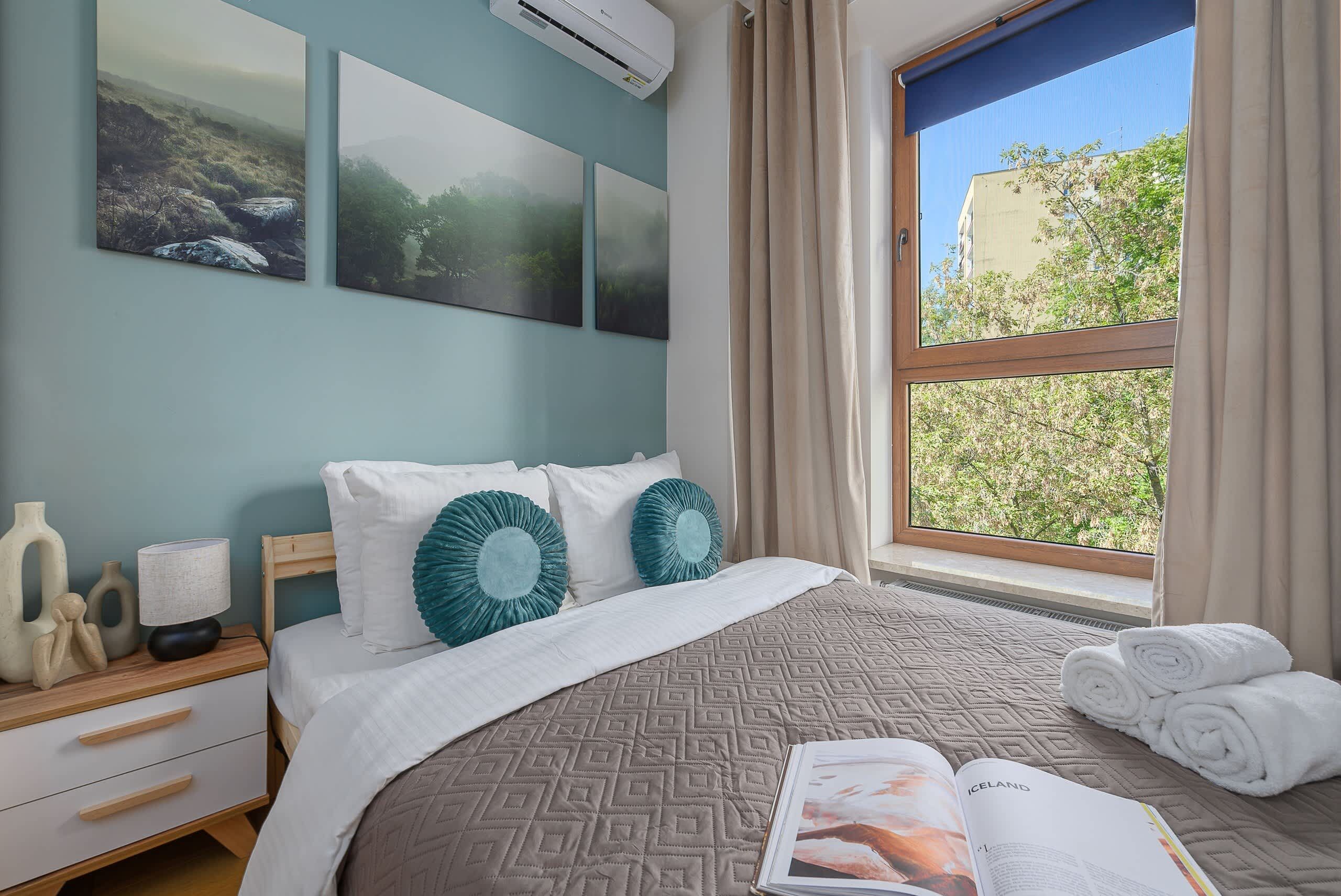 A bedroom with a bed, a nightstand, a window with curtains, and artwork on the wall.