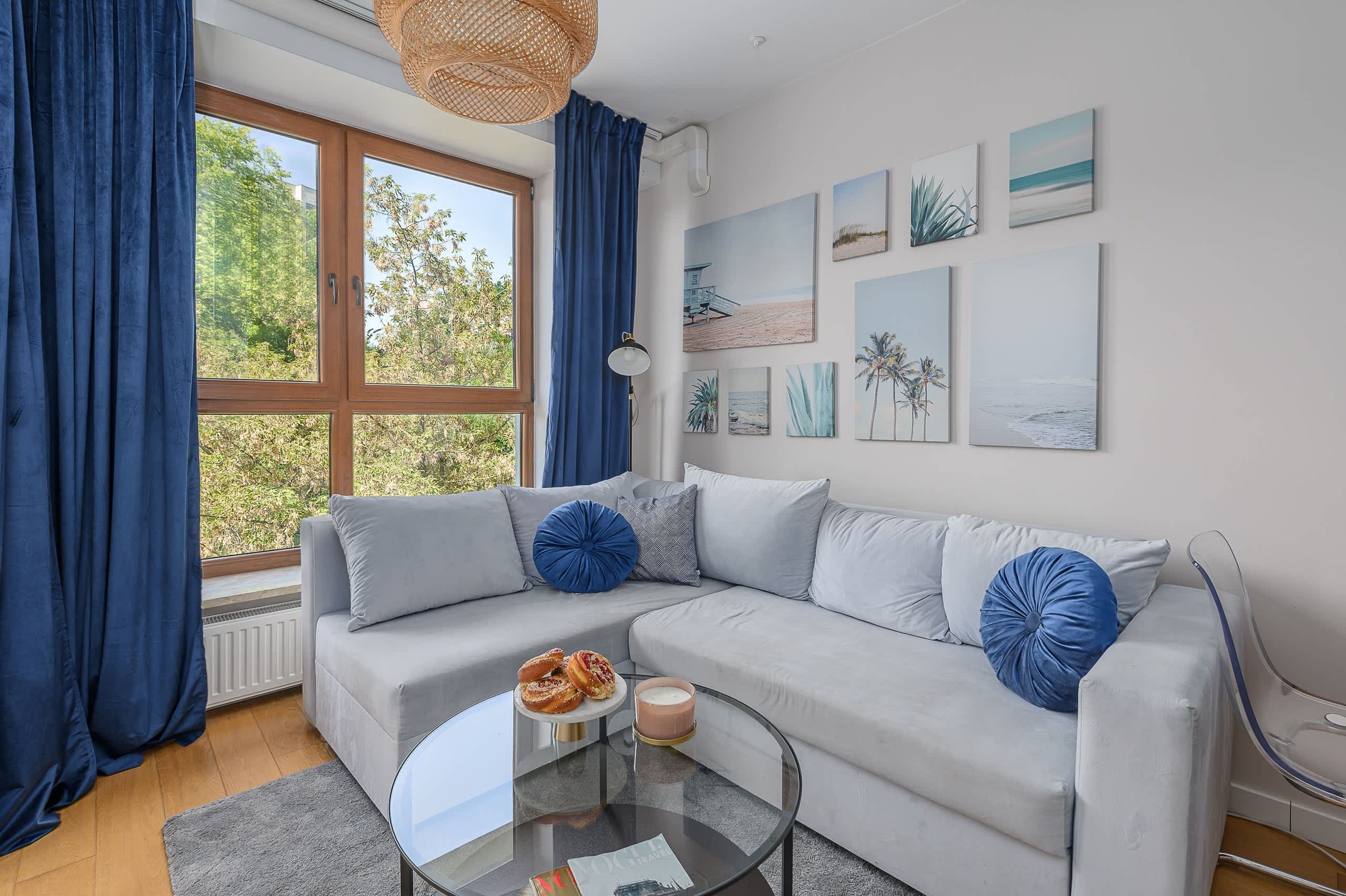 A modern living room with a grey sofa, blue cushions, a glass coffee table with pastries, and beach-themed wall art.