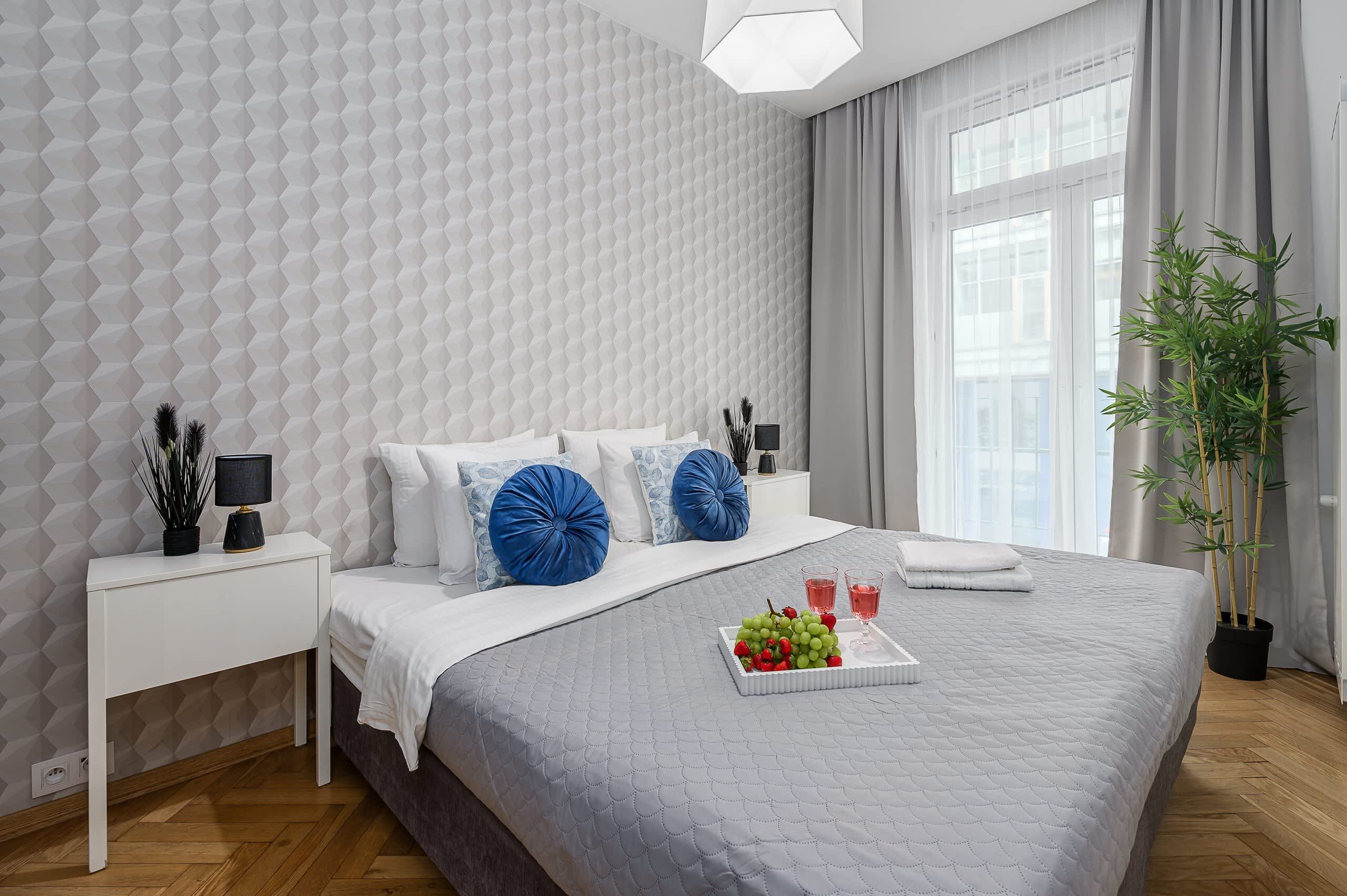 A modern bedroom with a geometric patterned wall, a bed with white bedding and blue accent pillows, a bedside table with a lamp, a tray with fruit and glasses, and a large window with curtains.