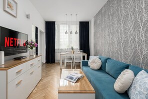 A modern living room with a flat-screen TV, a coffee table with a magazine, a blue sofa, and a dining area with a white table and chairs.