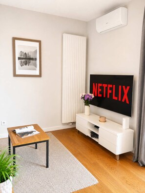 A modern living room with a flat-screen TV displaying 'NETFLIX,' a framed picture on the wall, a white radiator, and a small table with a magazine. - Lumina 3-room Apart w/ Parking & AC & Balcony (Warszawa)