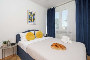 A hotel room with a bed, a bedside table with a lamp, a potted plant, a framed picture on the wall, and a window with curtains. - Lumina 3-room Apart w/ Balcony near Park & Metro (Warszawa)