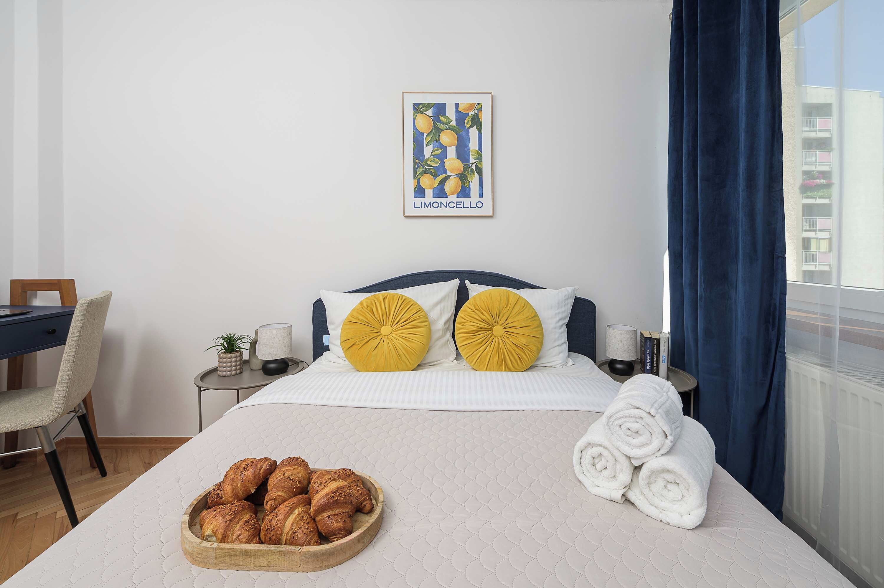 Habitació d'hotel amb llit, una taula de nit, una cadira, finestra amb cortines, una obra d'art a la paret i un safata amb pastissos.
