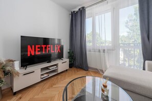 Ett modernt vardagsrum med en platt-TV som visar "NETFLIX" på skärmen, ett soffbord i glas, en soffa och utsikt över naturen genom stora fönster.