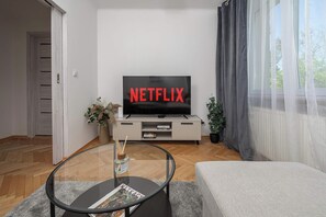 Ett modernt vardagsrum med en platt-TV som är monterad på väggen, ett glasbord med tidningar, en soffa och en krukväxt.