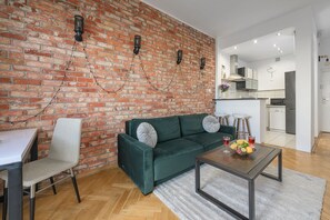 Ein Wohnzimmer mit Backsteinwand, einem grünen Sofa, einem Esstisch mit Stühlen und einer Küche im Hintergrund.