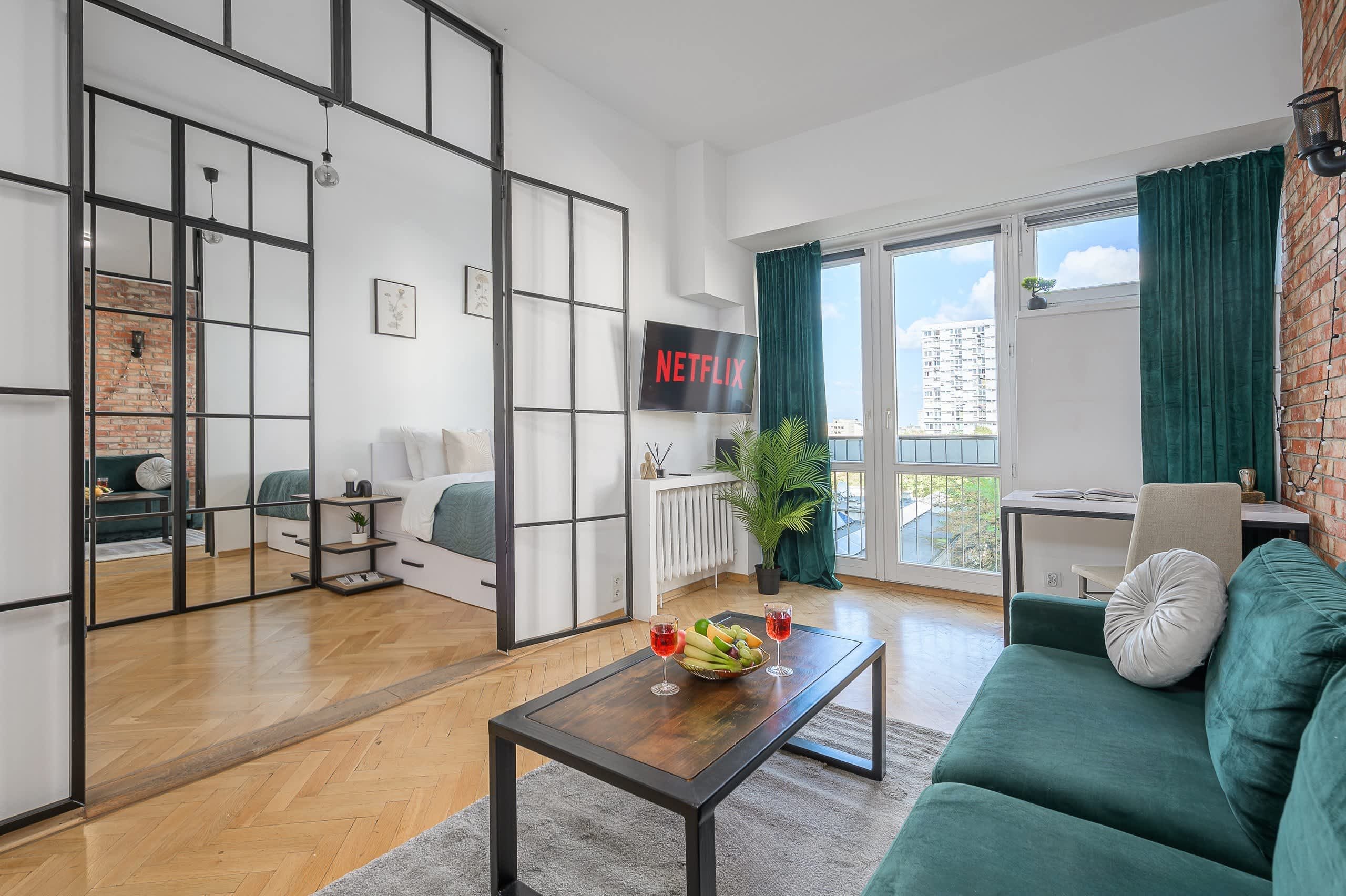A modern living room with a sofa, a coffee table with fruit and drinks, a TV displaying Netflix, and a view of a building outside.