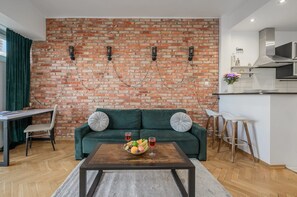 Een moderne woonkamer met een bakstenen muur, een groene bank, een houten salontafel met fruit en een keukenhoek met een roestvrijstalen afzuigkap.
