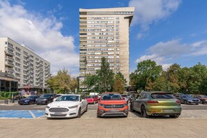 A parking lot with several cars, a tall residential building, and a smaller building with signage. - Lumina 3-room Apart w/ Balkony in the Center (Warszawa)