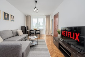 A modern living room with a grey sofa, a coffee table, a TV with a Netflix logo, and a dining area with a table and chairs. - Lumina 3-room Apart w/ Balcony near ZOO (Warszawa)