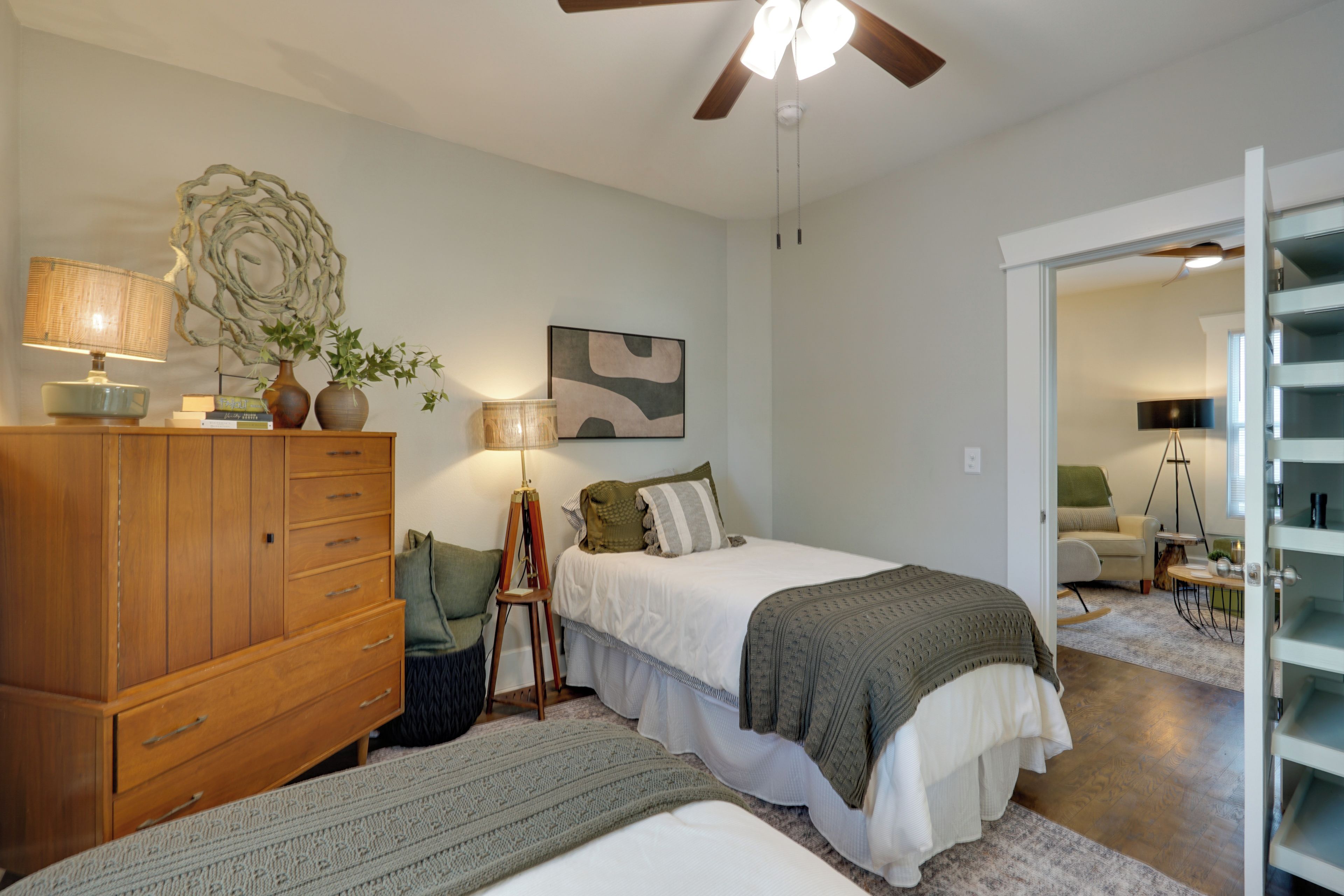 A bedroom with a wooden dresser, a standing lamp, a bed with a green blanket, and a ceiling fan.