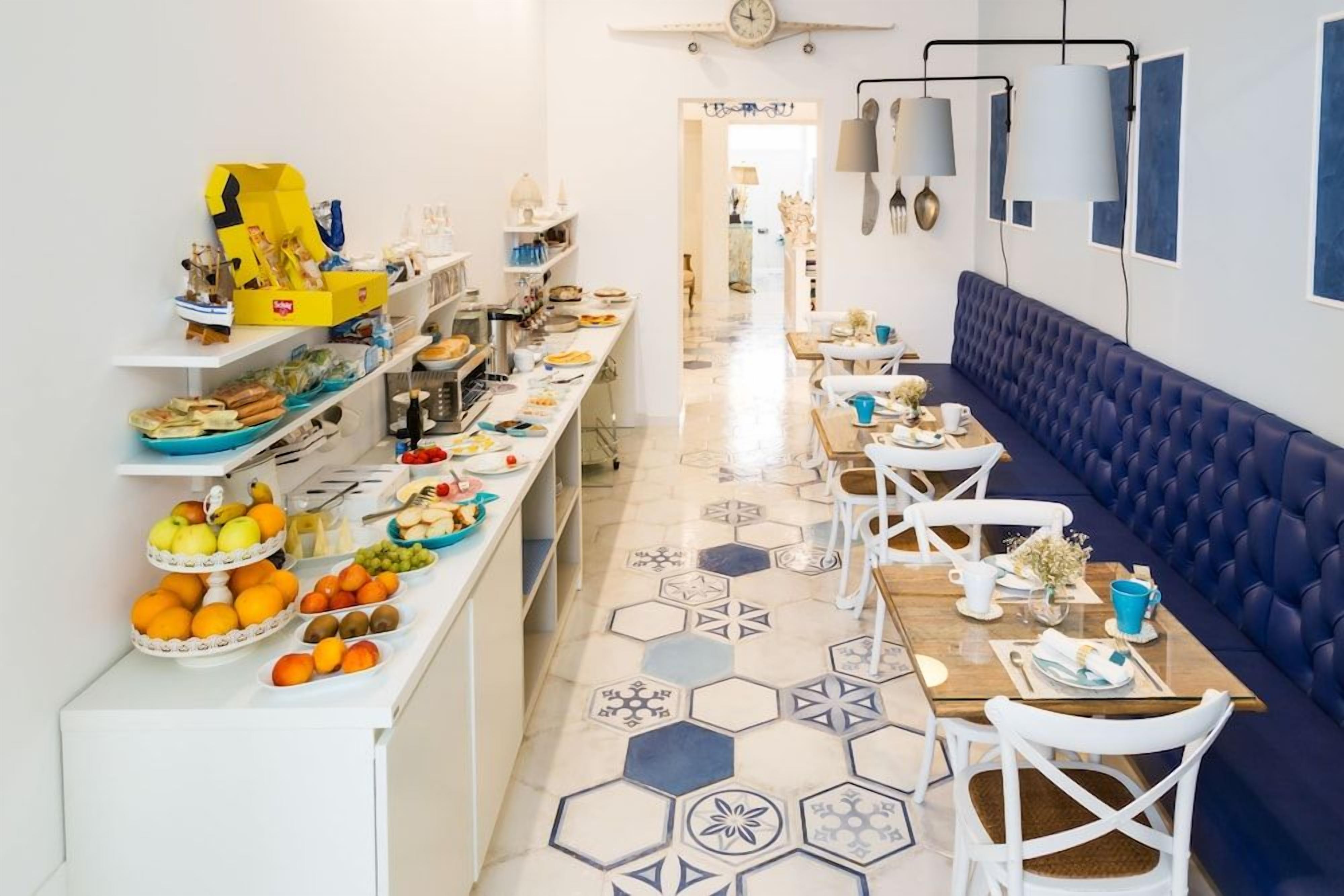 A long dining area with a blue cushioned bench, white chairs, and a variety of food items on the counter.