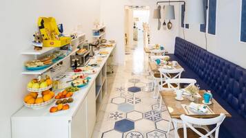 A long dining area with a blue cushioned bench, white chairs, and a variety of food items on the counter.