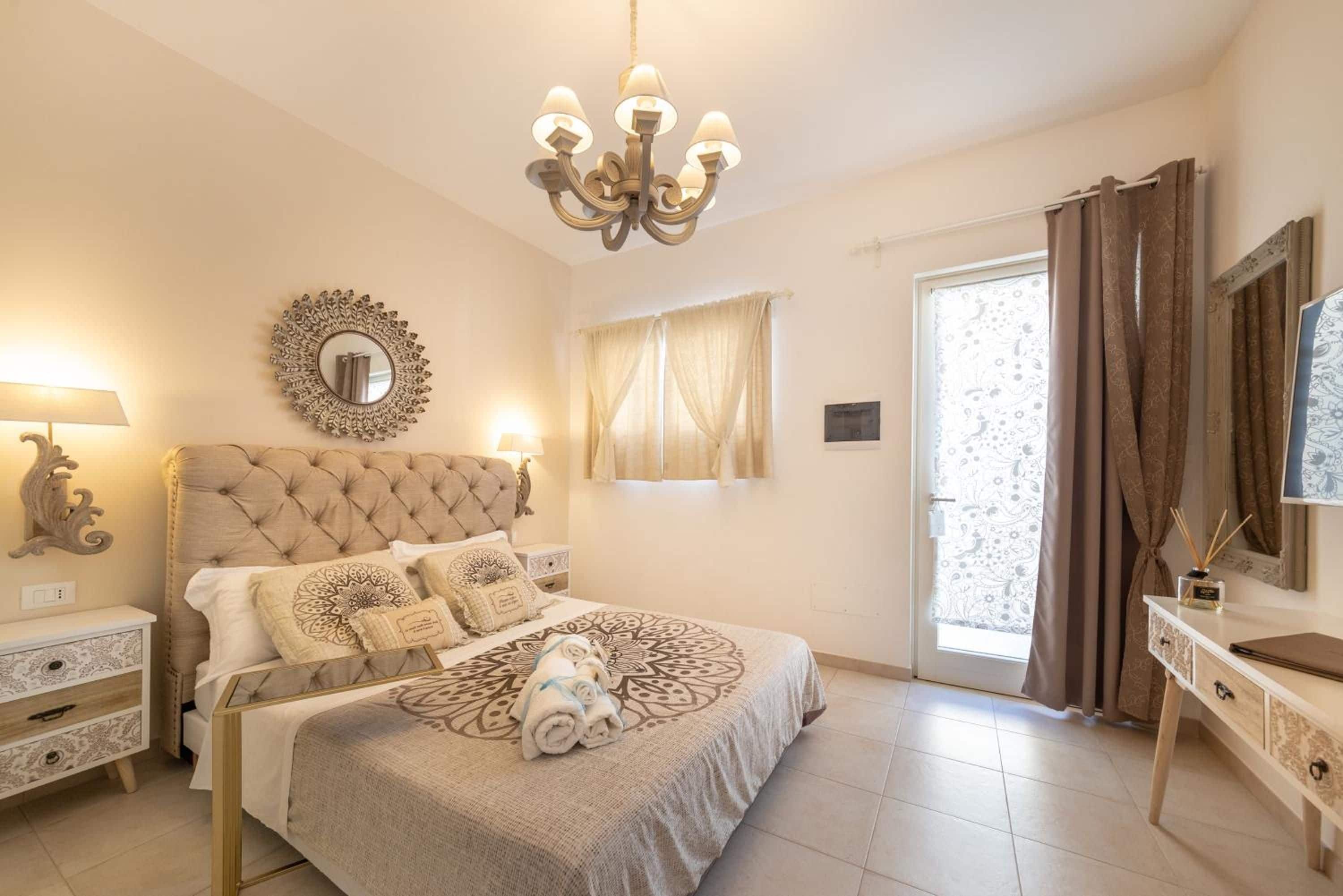 A bedroom with a tufted headboard, a chandelier, a mirror, a bedside table, a dresser, and a window with curtains.