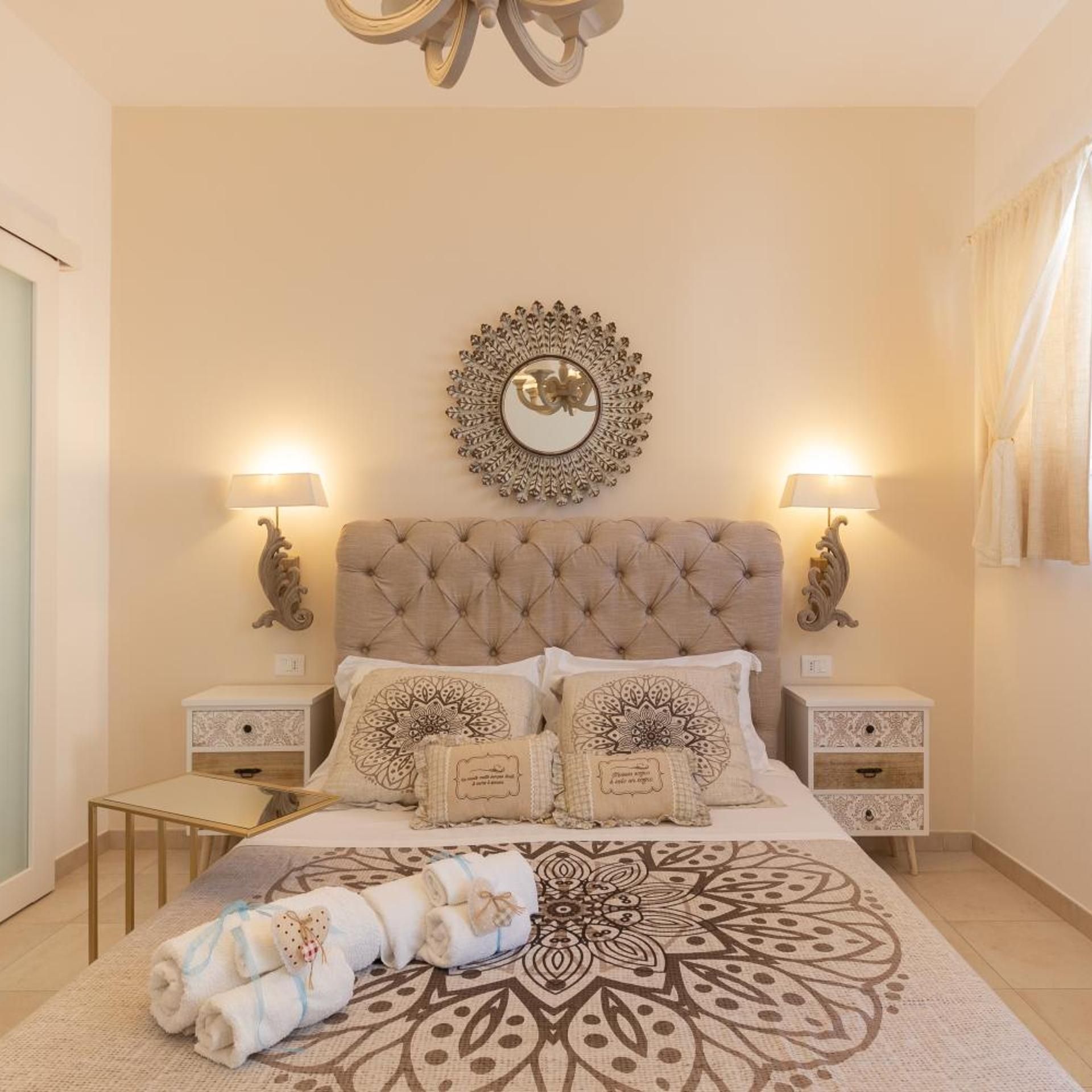 Un dormitorio con cabecera capitonada, un espejo decorativo, dos lámparas de noche, una colcha estampada y toallas enrolladas sobre una mesita de noche.
