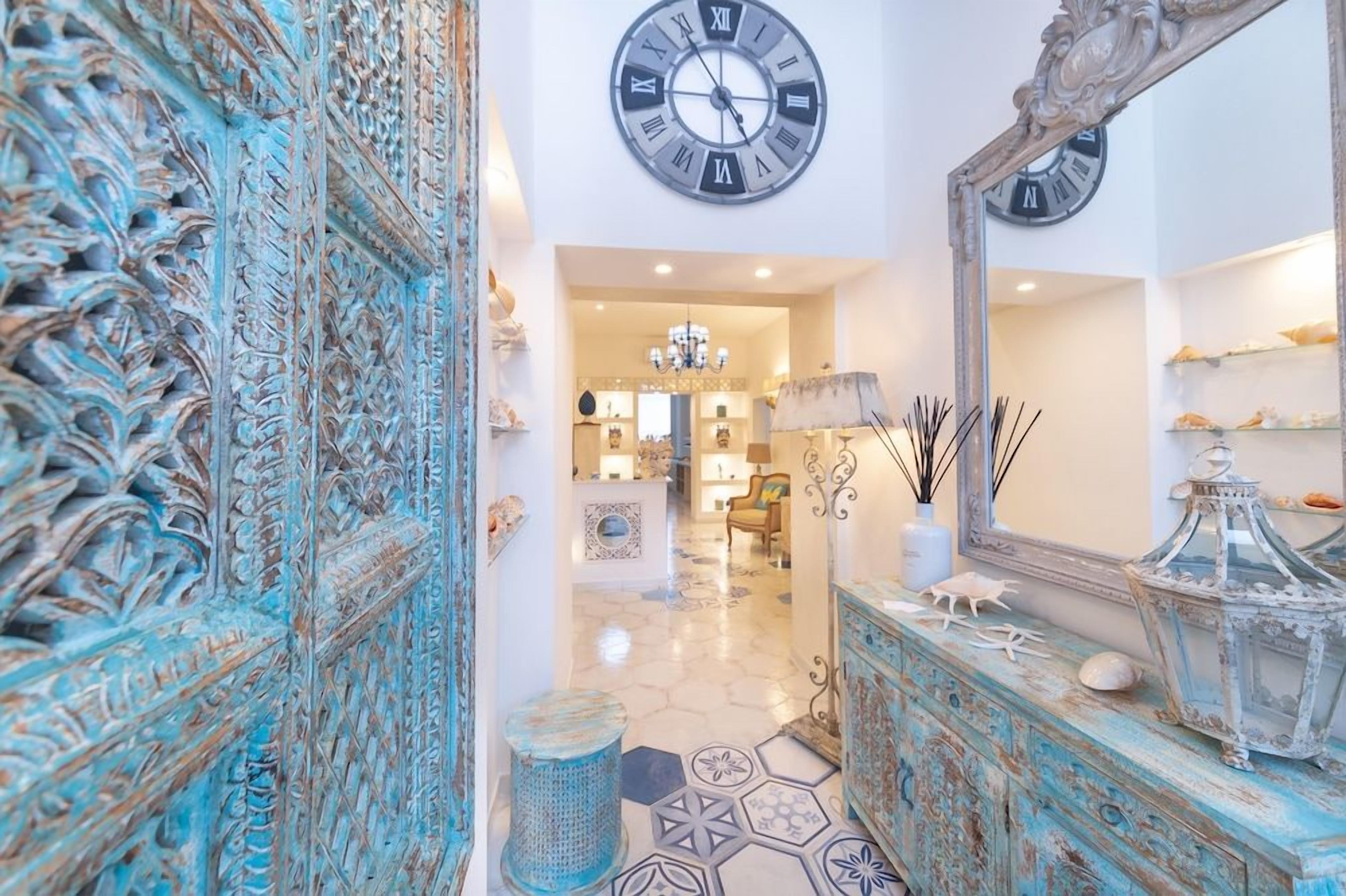 A hallway with a decorative blue door, a large wall clock, and a mirror reflecting a chandelier.