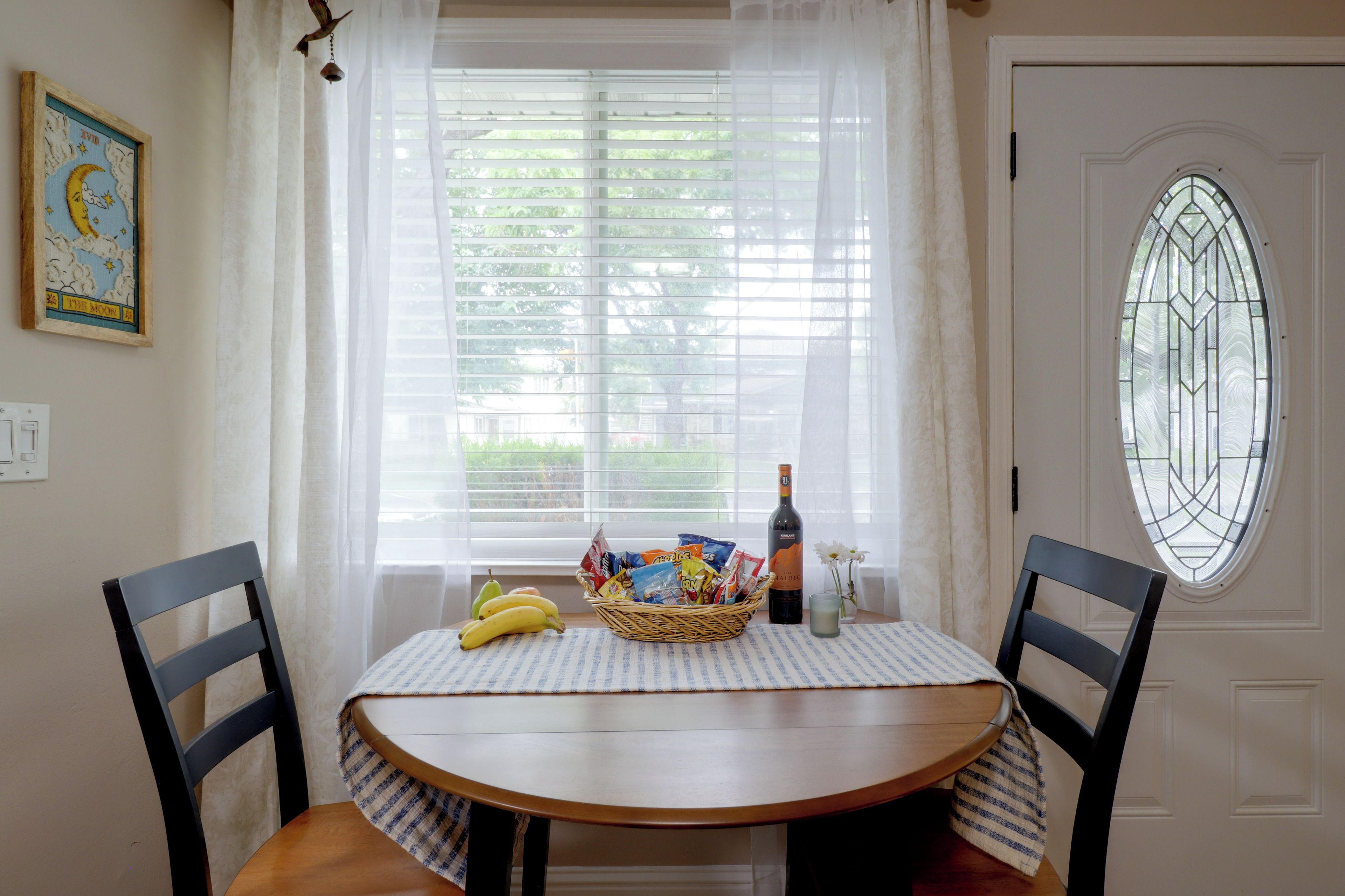 Un área de comedor con una mesa preparada para dos, una cesta de aperitivos, una botella de vino y un florero con flores.