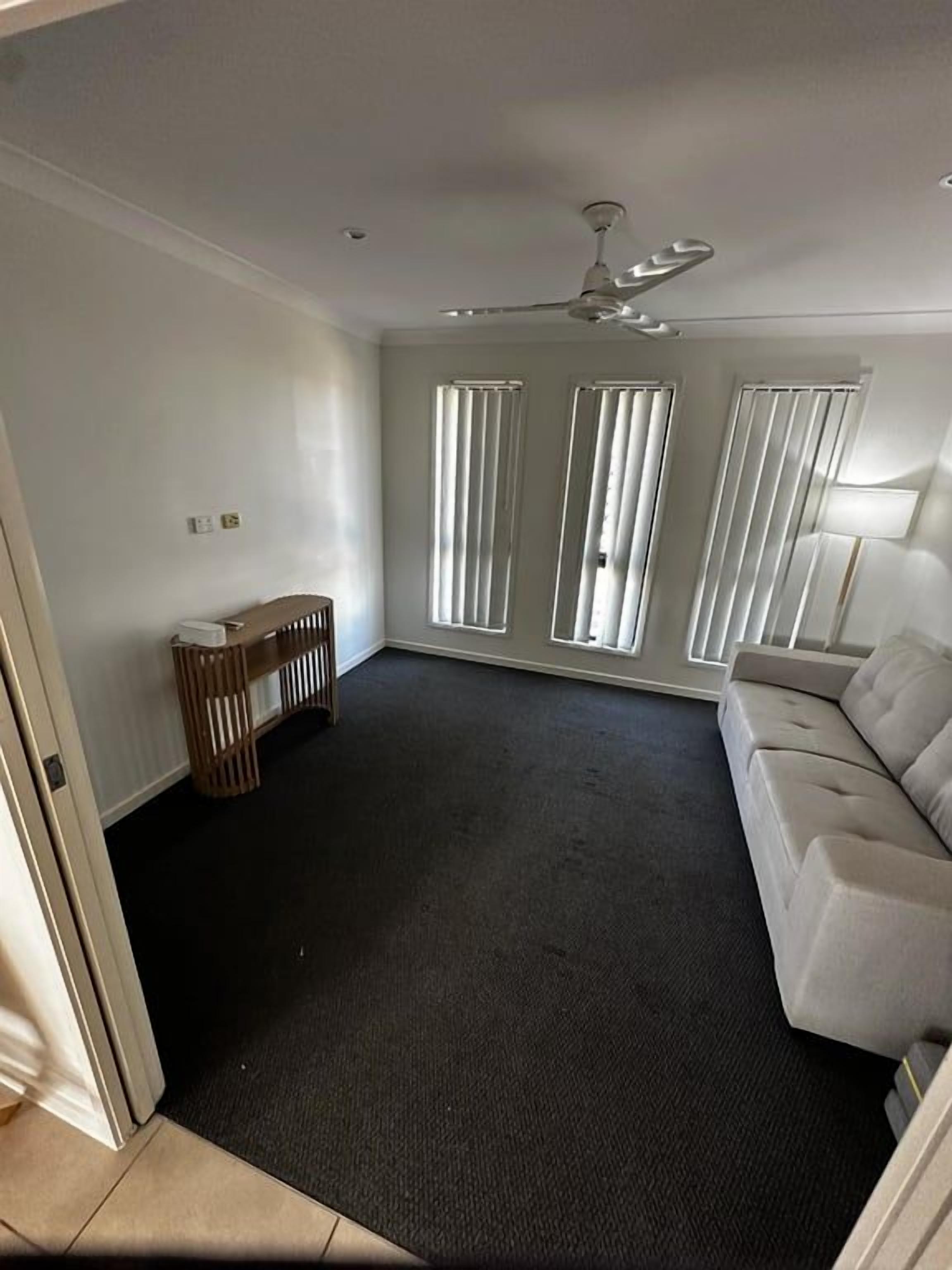 A room with a dark carpet, a white sofa, a ceiling fan, and a window with blinds.