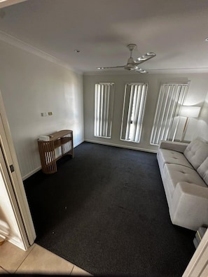 A room with a dark carpet, a white sofa, a ceiling fan, and a window with blinds. - WHITE + HAVEN - Entire home (Mackay)