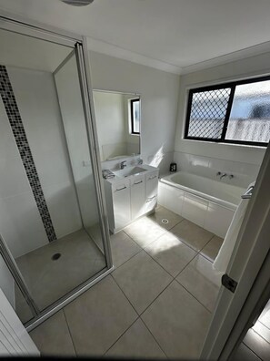A modern bathroom with a glass shower enclosure, a bathtub, and a window providing natural light. - WHITE + HAVEN - Entire home (Mackay)