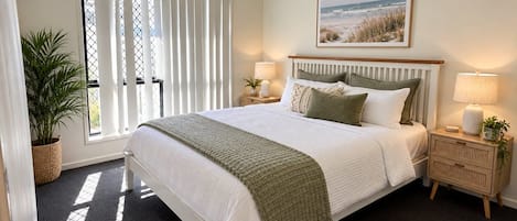 Une chambre à coucher comprenant un lit, un ventilateur de plafond, une fenêtre avec des stores, une table de chevet avec une lampe et un tableau accroché au mur.