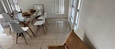 A dining area with a glass table and chairs, a wooden bench, and large windows with blinds.