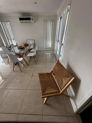 A dining area with a glass table and chairs, a wooden bench, and large windows with blinds. - WHITE + HAVEN - Entire home (Mackay)